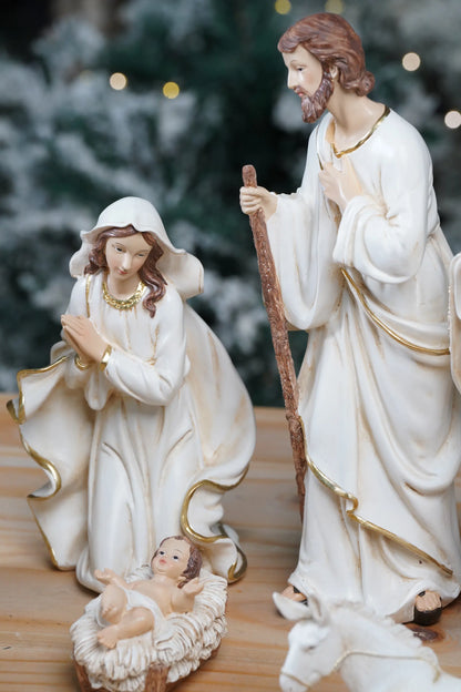 White and gold nativity Holy Family close-up — kneeling Mary, Joseph with staff, and Baby Jesus in manger against snowy Christmas tree background