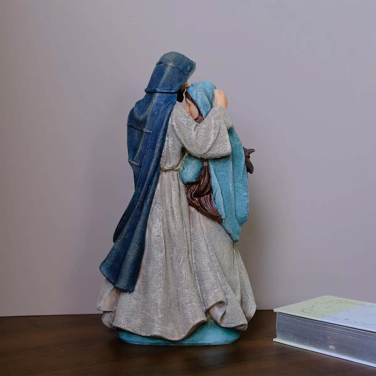 Holy Family statue back view on wood tabletop beside book — teal and blue hand-painted resin nativity figurine