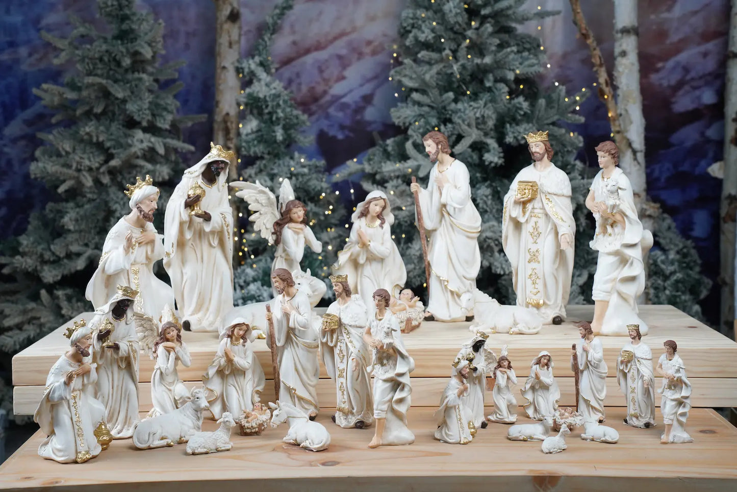 Multiple sizes of white and gold nativity sets displayed together on tiered wooden platform with winter tree backdrop