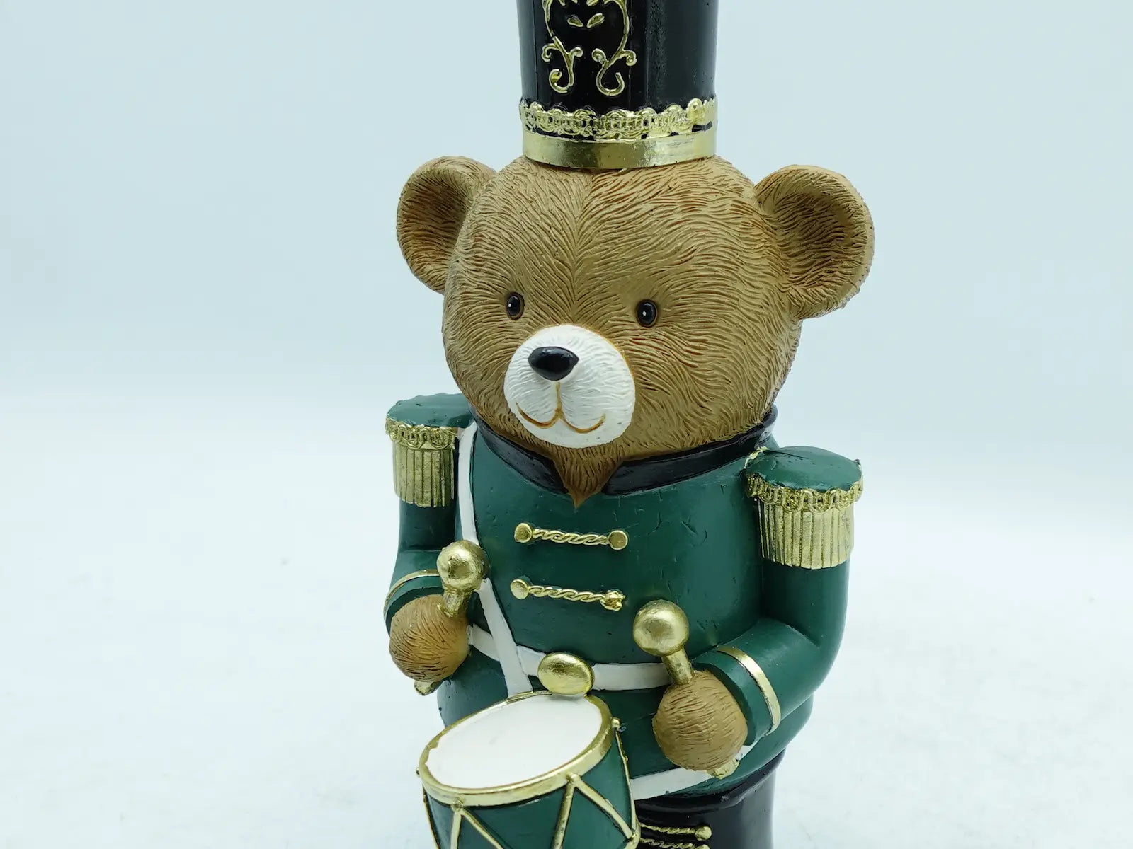 A teddy bear nutcracker figurine wearing a green uniform with gold trim and a black hat, holding a small drum against a plain background.