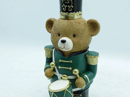 A teddy bear nutcracker figurine wearing a green uniform with gold trim and a black hat, holding a small drum against a plain background.