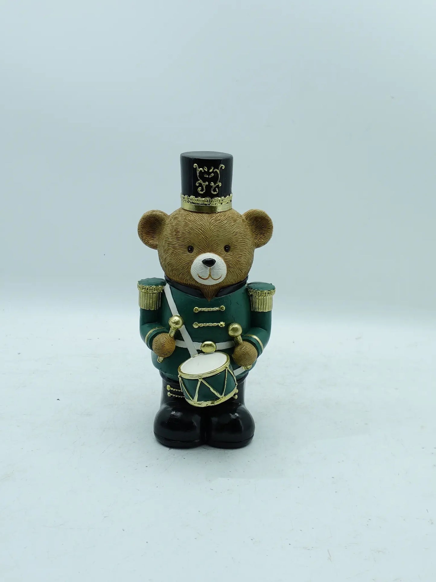 A Christmas teddy bear figurine dressed as a toy soldier, holding a drum, photographed against a plain light gray background.