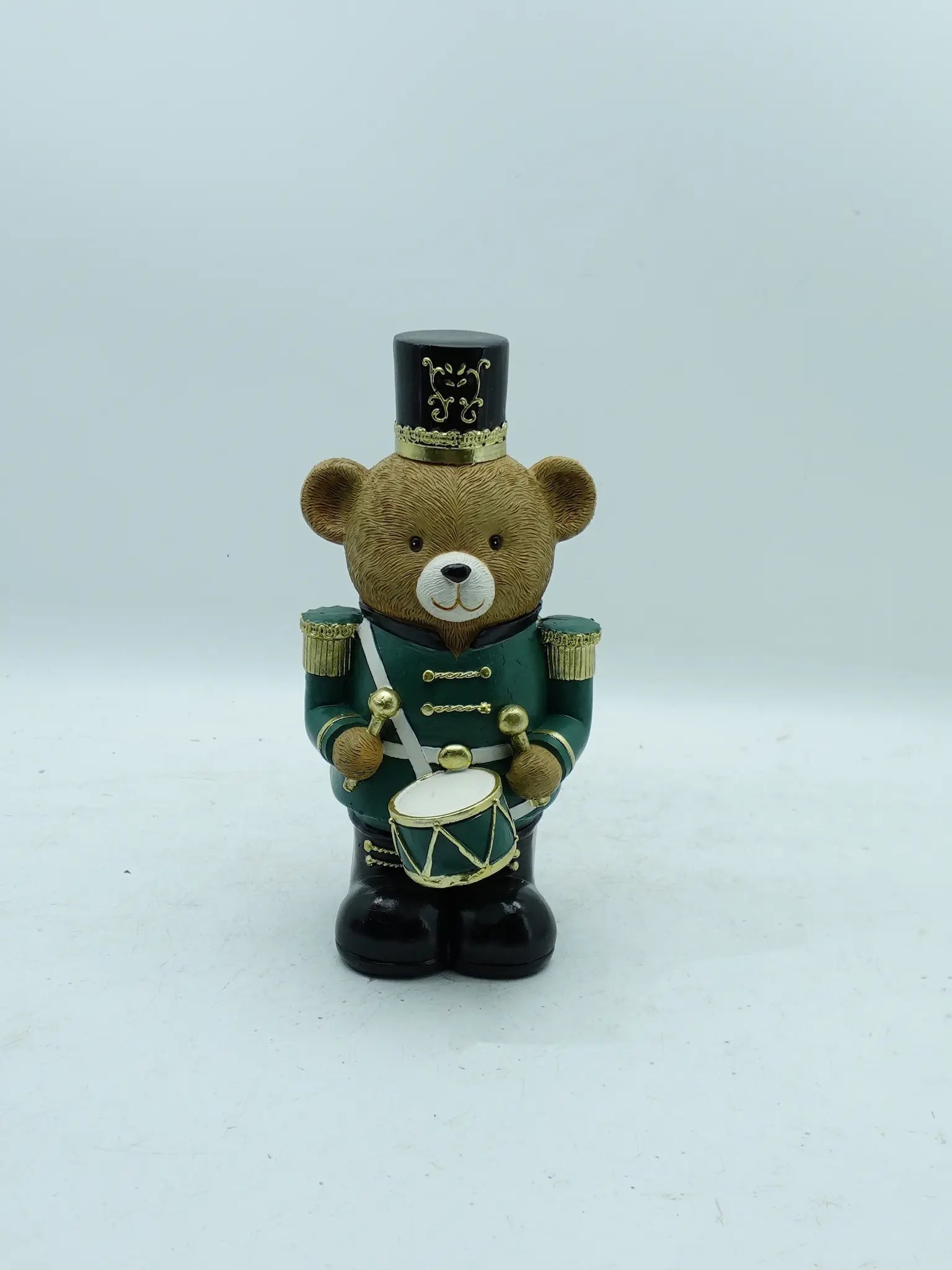 A Christmas teddy bear figurine dressed as a toy soldier, holding a drum, photographed against a plain light gray background.
