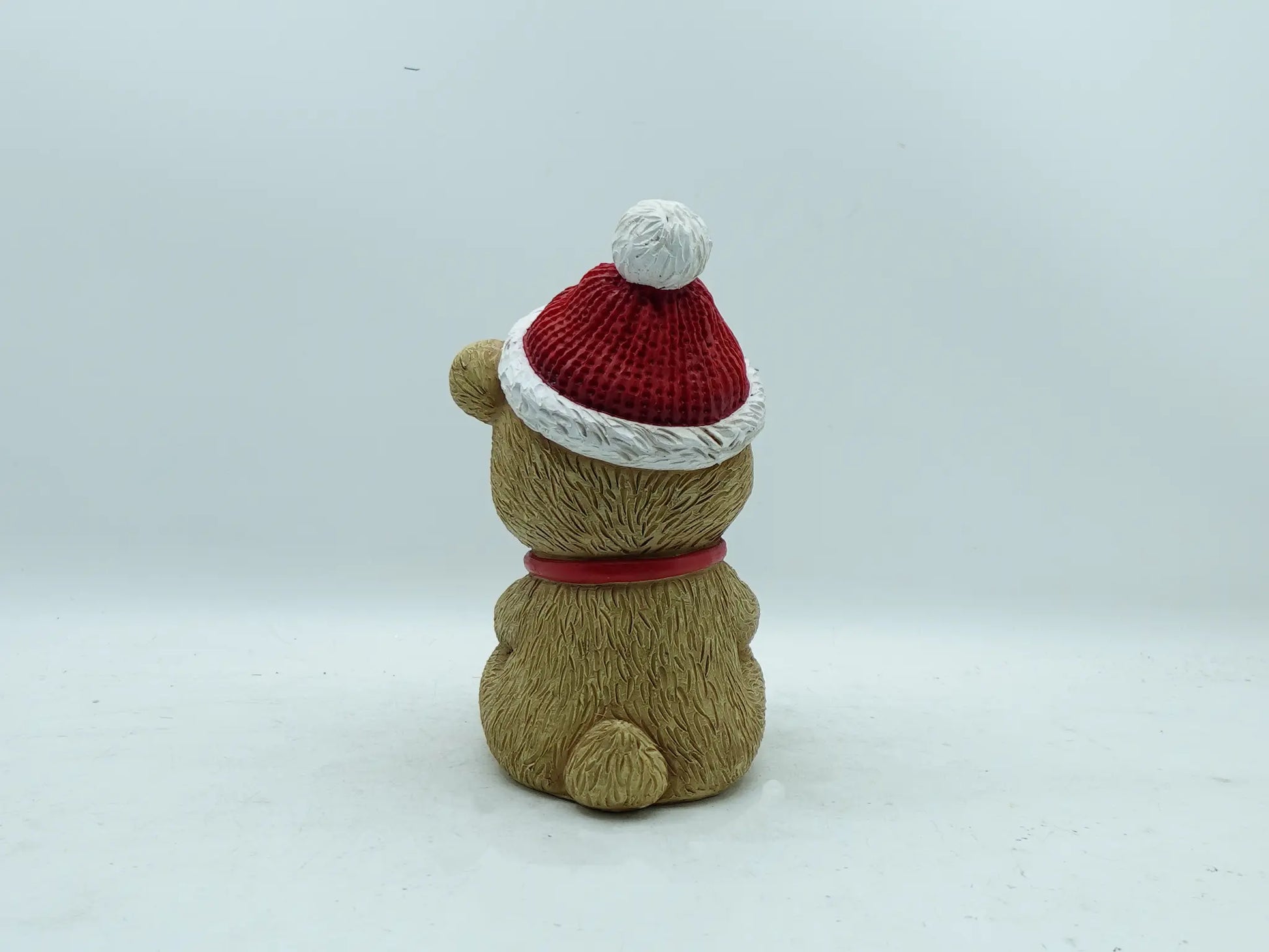 the teddy bear figurine showing the textured fur, red collar, and pom-pom hat from behind