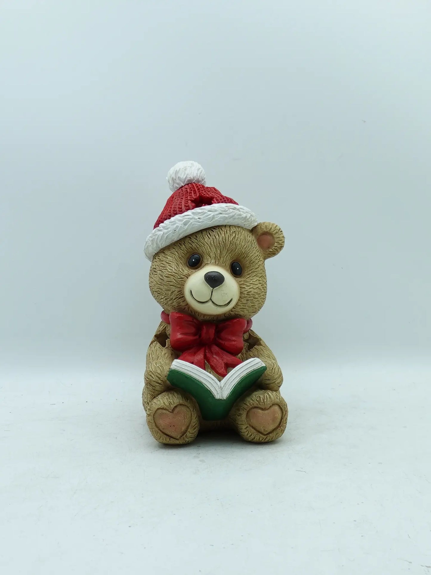 Hand-painted Christmas teddy bear figurine in red knit Santa hat with white pom-pom, large red bow, holding an open green storybook, front view