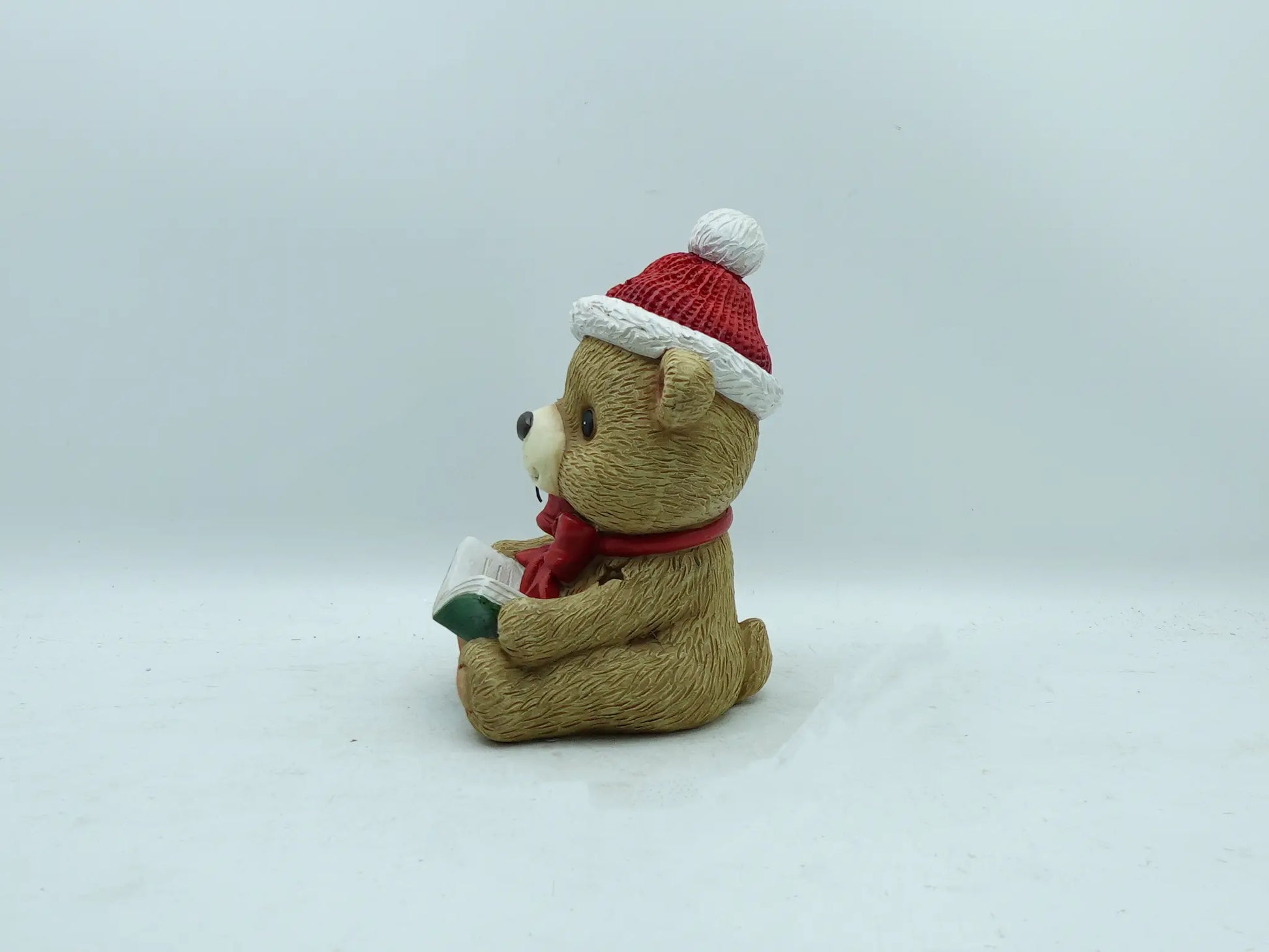 A beige teddy bear figurine wearing a Santa hat and red scarf, seated and holding a book.