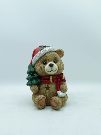A holiday teddy bear figurine carrying a green Christmas tree, photographed against a plain light gray background.