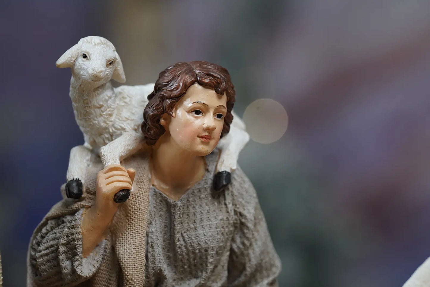 Young shepherd nativity figurine close-up — carrying lamb on shoulder in grey linen-texture robe with gold accent