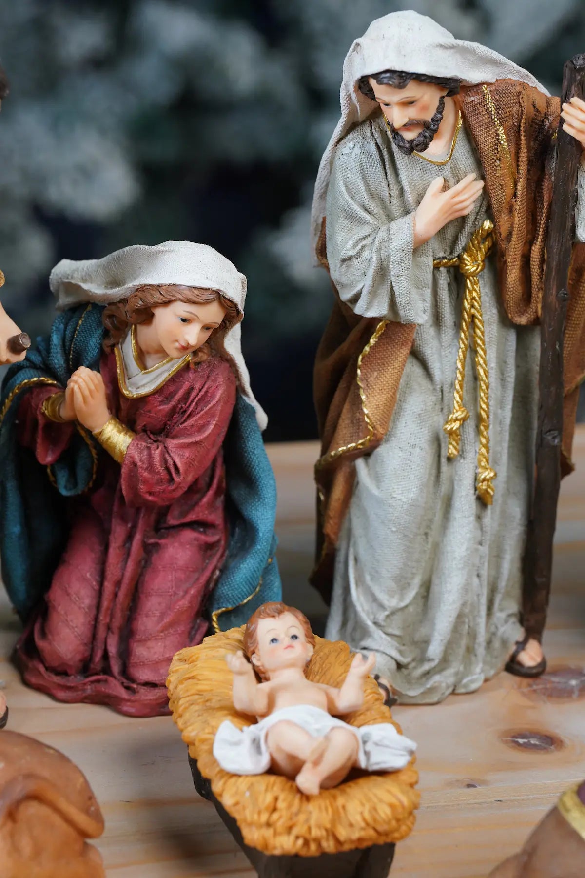 Holy Family close-up — kneeling Mary in crimson robe with teal mantle and standing Joseph with hand on chest beside straw manger