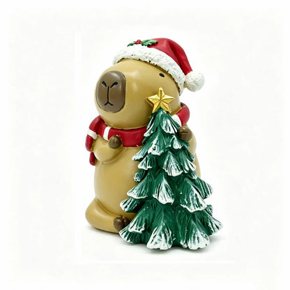 Hand-painted Christmas capybara figurine wearing Santa hat and red-white striped scarf, hugging a snow-dusted mini tree with gold star on white background