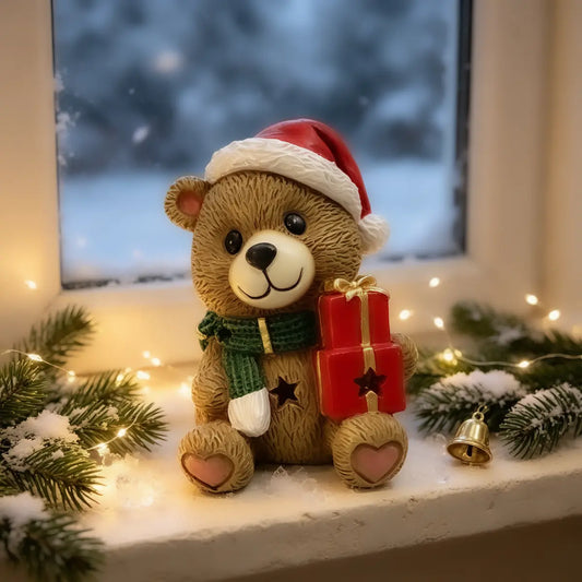 A teddy bear figurine wearing a red Santa hat and green scarf, sitting on a snowy windowsill and holding a red gift box with a gold bow.