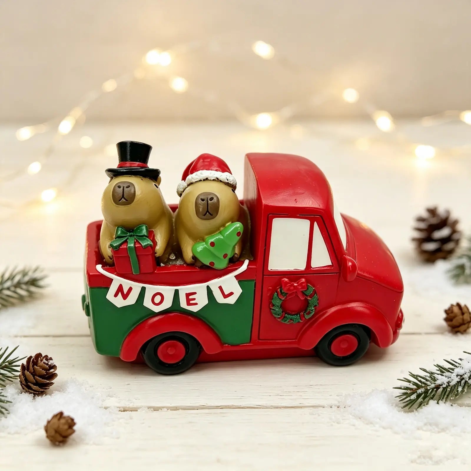 A festive resin holiday truck figurine carrying two capybaras, set on a snowy white surface with pinecones and warm string lights in the background.