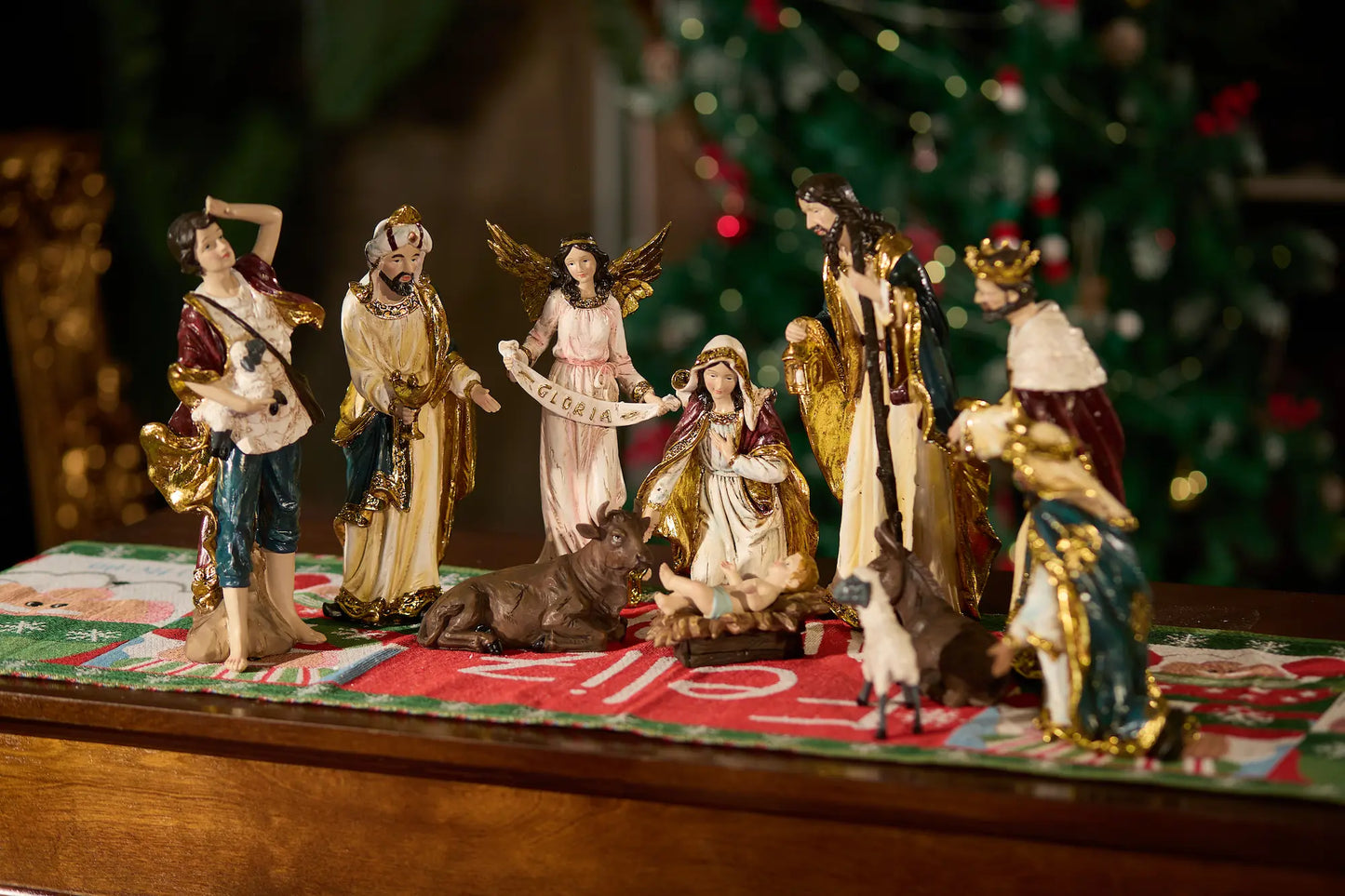 Complete nativity scene set with Gloria angel displayed on Christmas table runner with decorated tree in background