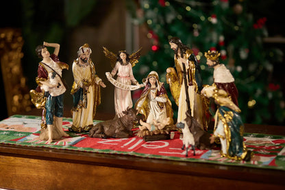 Complete nativity scene set with Gloria angel displayed on Christmas table runner with decorated tree in background