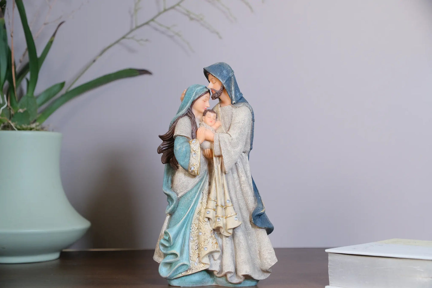 Holy Family statue on tabletop beside potted plant and books — hand-painted resin Mary and Joseph figurine with Baby Jesus