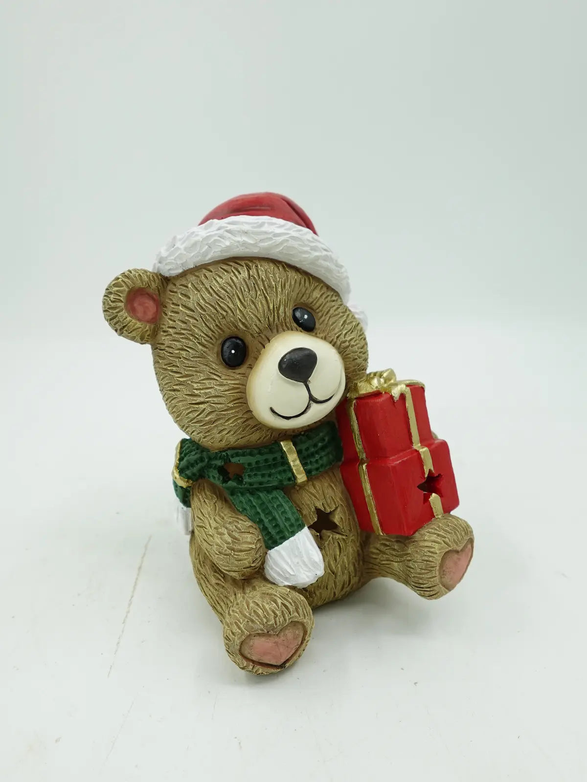 A Christmas teddy bear figurine, holding a red gift box with gold trim, set against a plain white background.