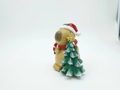 A capybara figurine wearing a red Santa hat and red scarf, holding a green Christmas tree with a gold star on a white background.