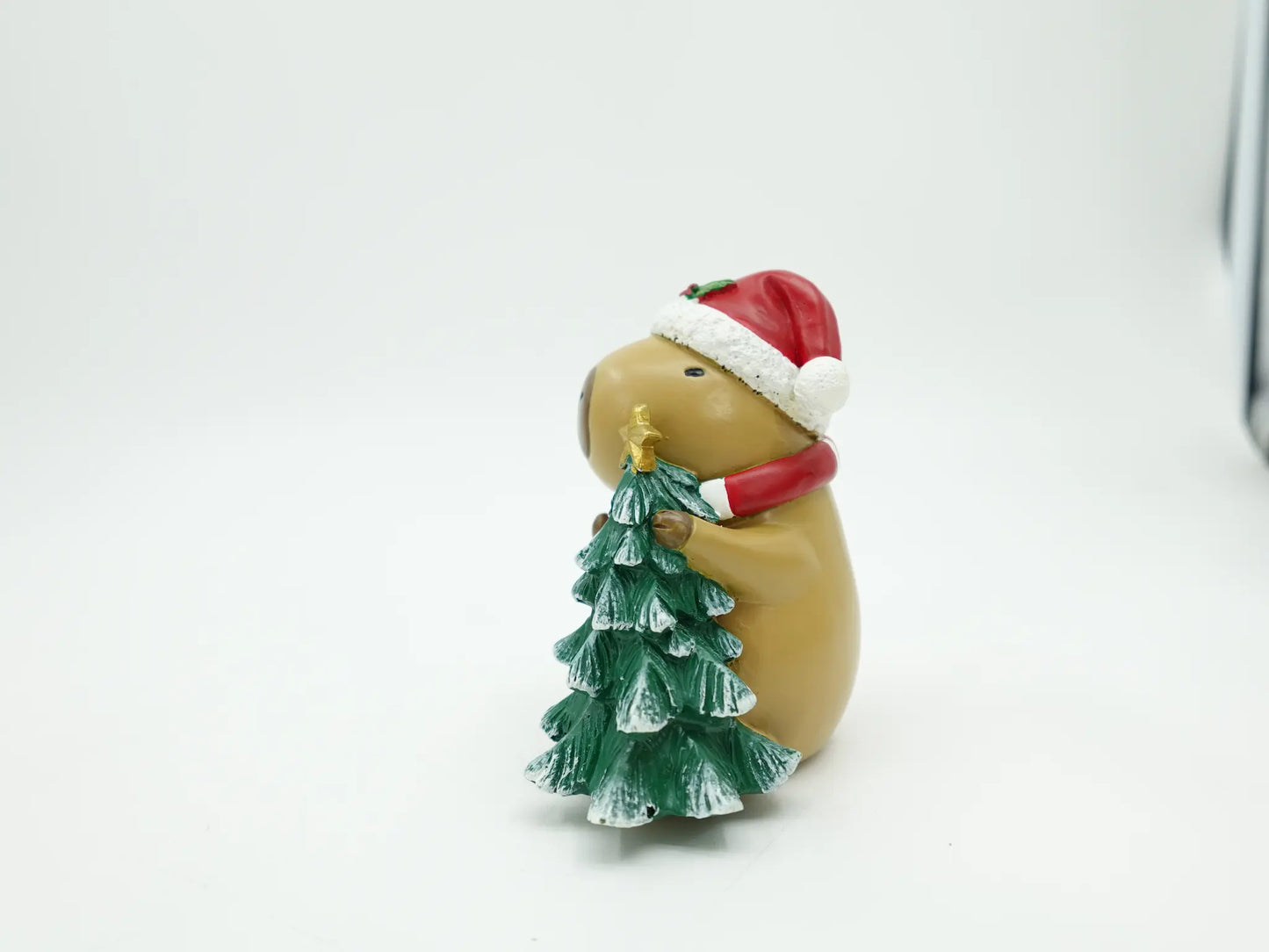 the capybara figurine hugging the snow-frosted mini Christmas tree with gold star topper, alternate side view 