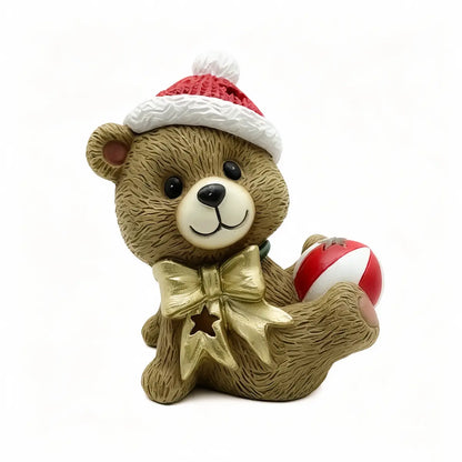 A brown teddy bear figurine wearing a red and white Santa hat, a gold bow tie, and holding a red and white ball, set against a plain white background.
