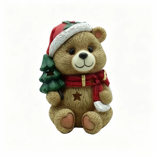 A teddy bear Christmas ornament figurine holding a small green Christmas tree, wearing a Santa hat and a red scarf with gold stars.